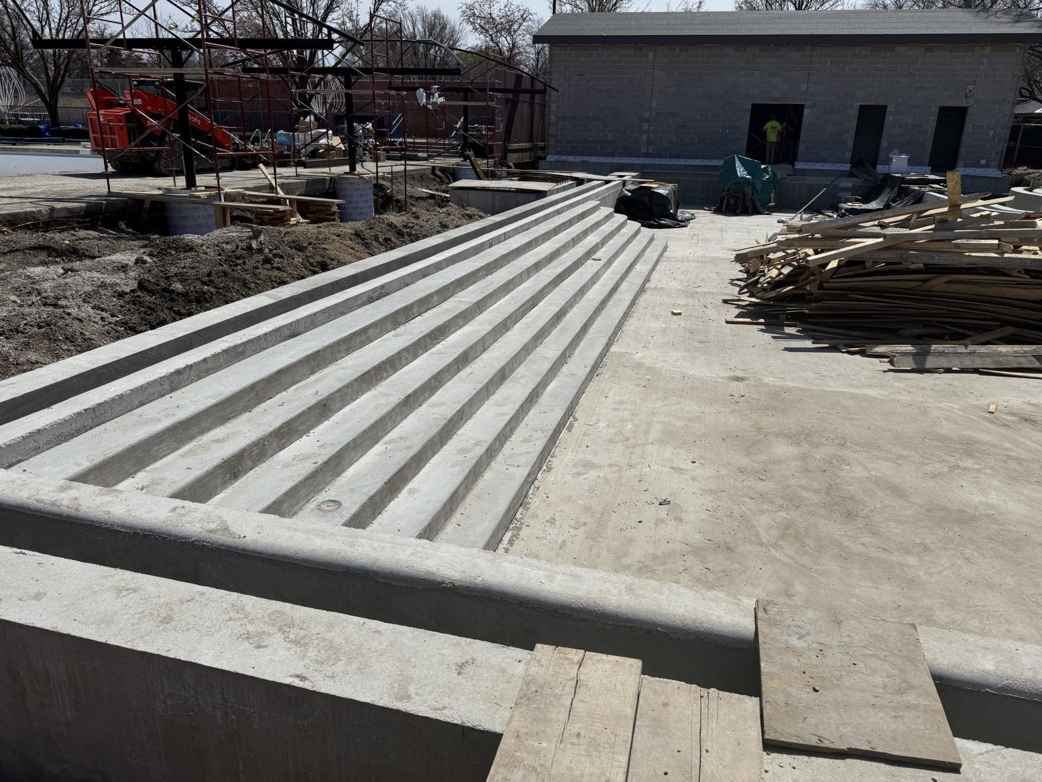 Newly poured concrete steps at Rehm Park & Pool construction site. A building and construction materials are visible in the