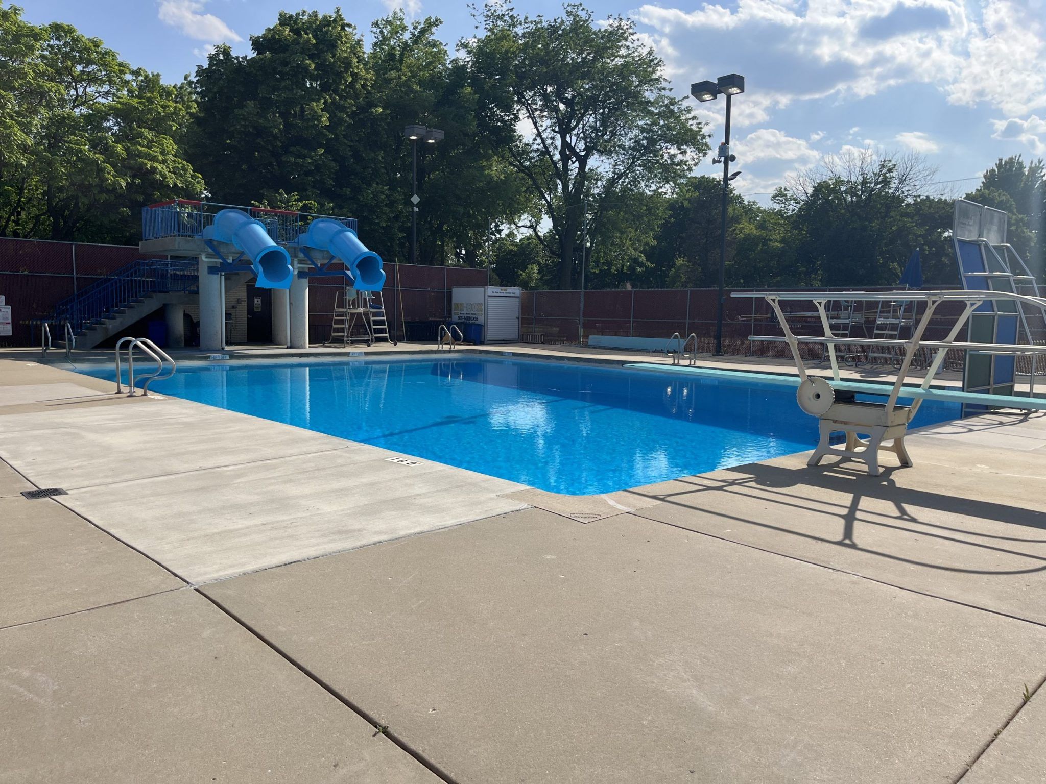 Rehm Park & Pool features a bright blue swimming pool with two blue water slides, a diving board, and surrounding concrete
