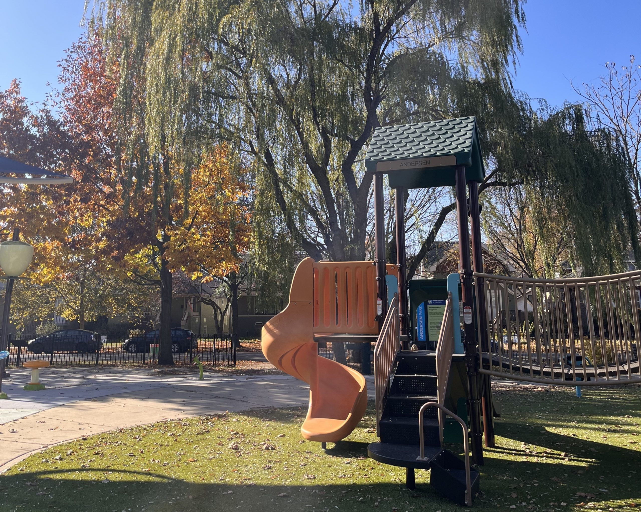 Playground structure with orange slide and stairs at Andersen Park, surrounded by trees with autumn foliage.