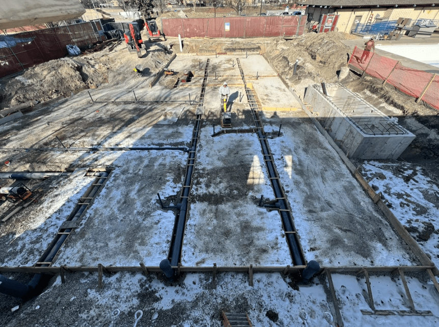 Construction site with concrete foundation, rebar, and plumbing pipes laid out. A worker operates a compactor.