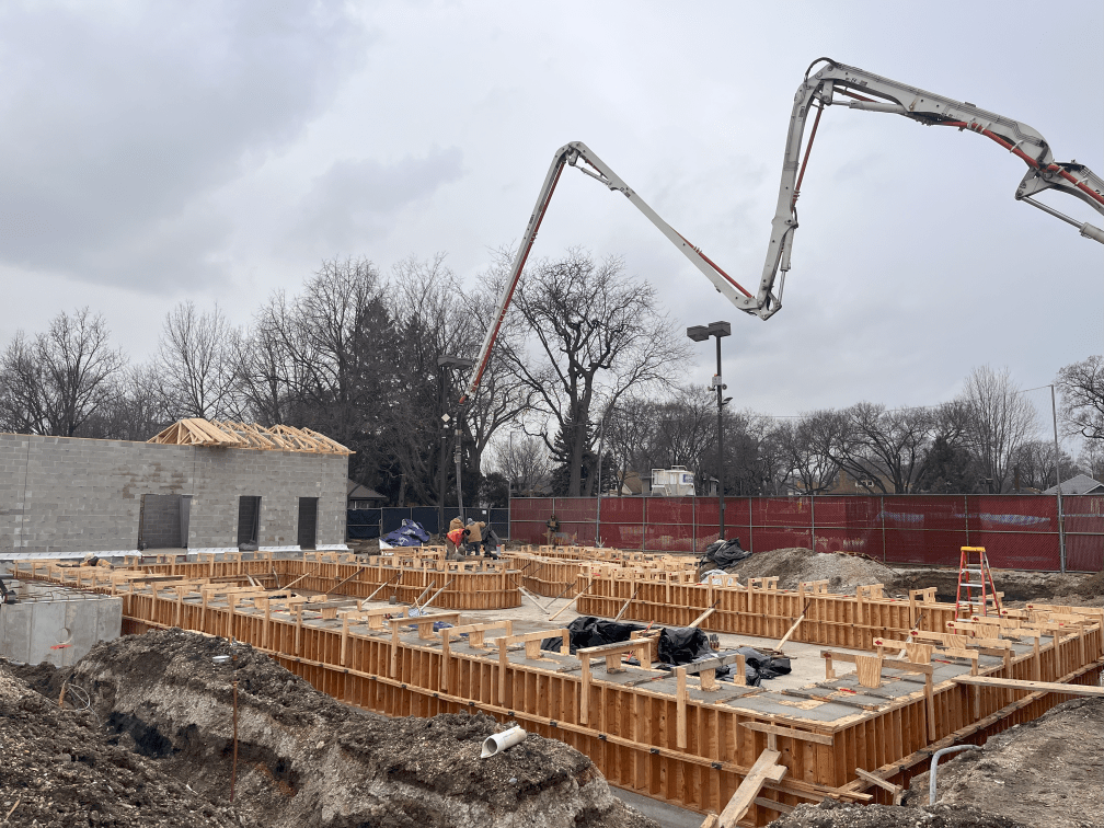 Construction of Rehm Park & Pool: concrete pump truck pouring foundation walls, with cinder block building in background.