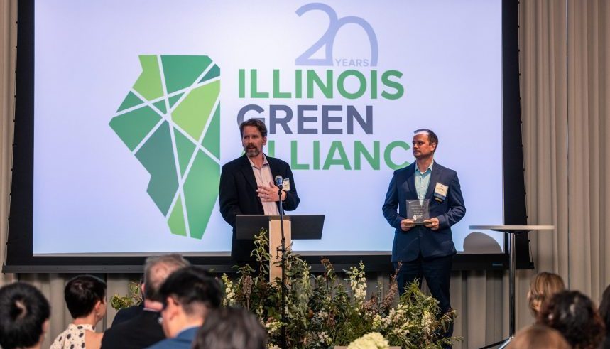 Two men stand before a screen displaying the Illinois Green Alliance logo and "20 Years." One man speaks at a podium, the