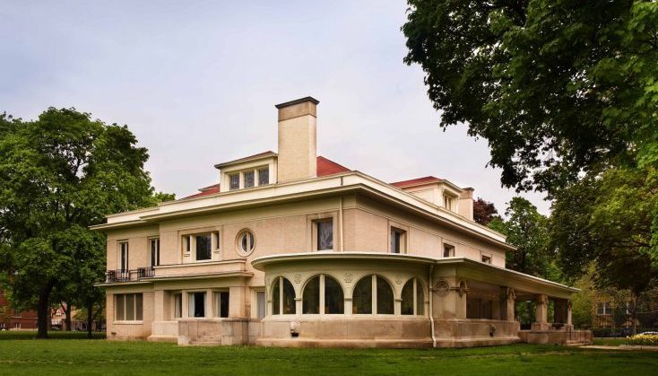 A large, light-colored historic house with a red roof, featuring a prominent chimney and a curved sunroom with arched