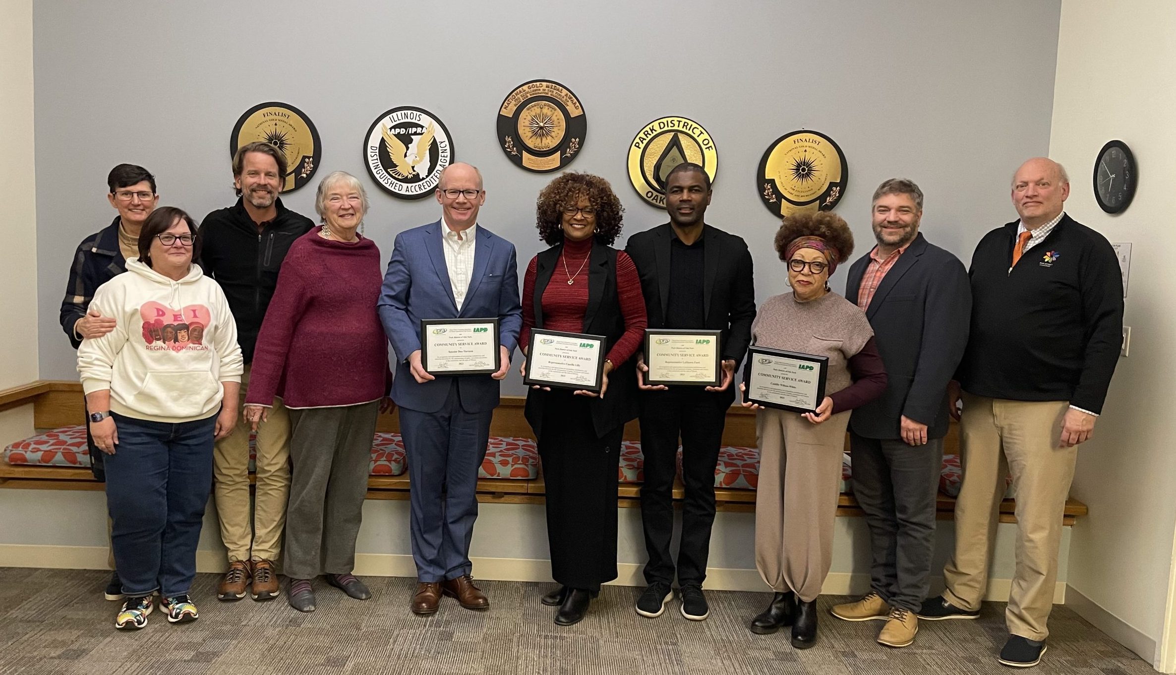 Group of ten people standing and smiling, holding awards. Several circular plaques are mounted on the wall behind them.