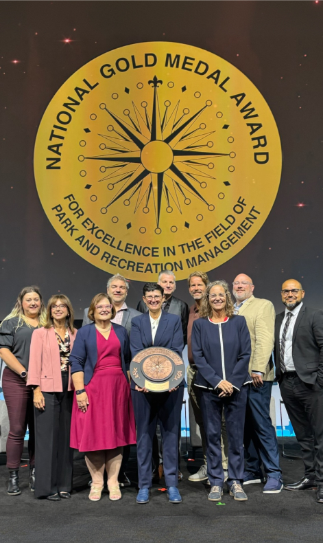 A group of ten people stand together, posing for a photo in front of a large golden "National Gold Medal Award" emblem.