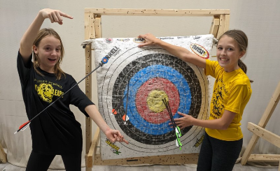 Two girls posing with an archery target. One girl in a black shirt points to an arrow in the target, the other in a yellow
