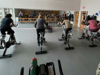 Several people are participating in a spin class, cycling on stationary bikes in a gym. A large mirror reflects more