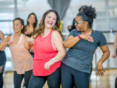 Two women laughing and dancing in a fitness class. Other participants are visible in the background.