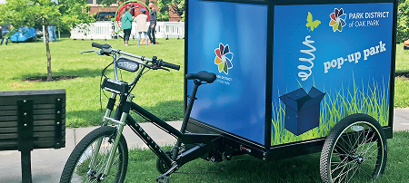 A three-wheeled bicycle pulling a cargo trailer branded with "Park District of Oak Park" and "pop-up park".