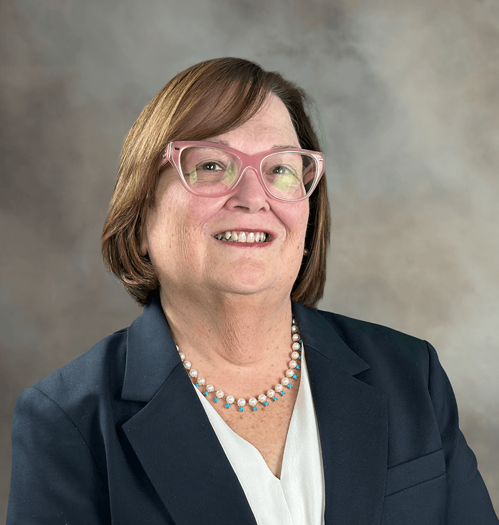 Portrait of a woman with short brown hair, pink cat-eye glasses, wearing a dark blazer and a pearl and turquoise necklace.