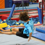 A young boy in a blue gymnastics outfit tumbles through the air over a blue mat, with foam pits and equipment in the