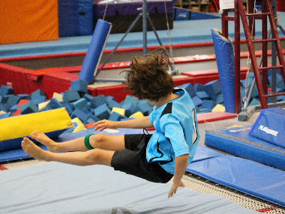 A young boy in a blue gymnastics outfit tumbles through the air over a blue mat, with foam pits and equipment in the