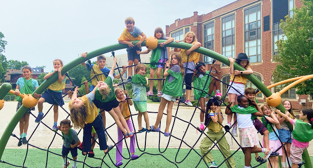 A diverse group of children in green and yellow shirts are playing on a large climbing net structure outside a brick school