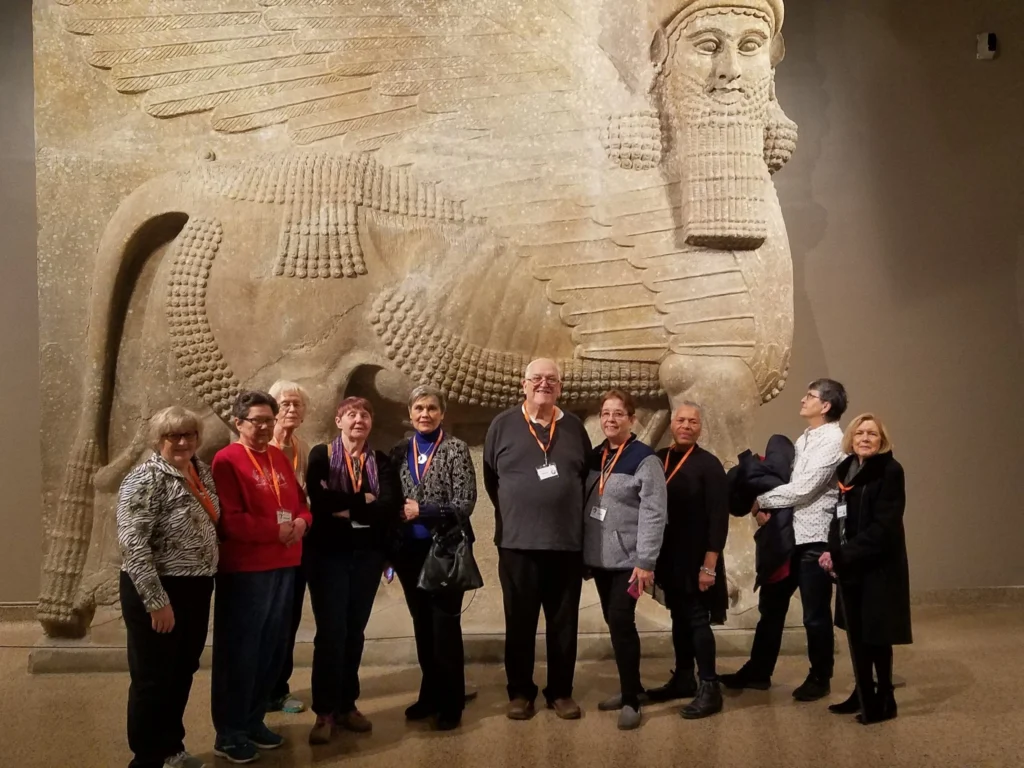 A group of ten people stand in front of a large, ancient stone sculpture of a winged bull with a human head.