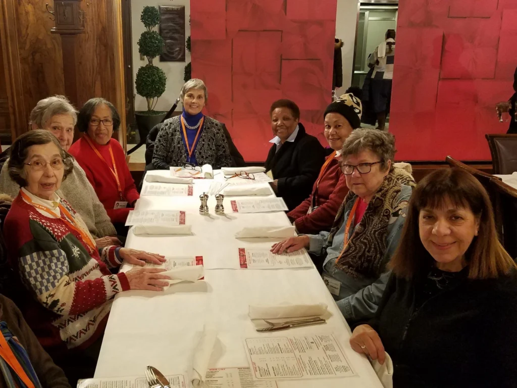 Group of eight women of various ages seated at a long table in a restaurant, smiling and looking towards the camera.