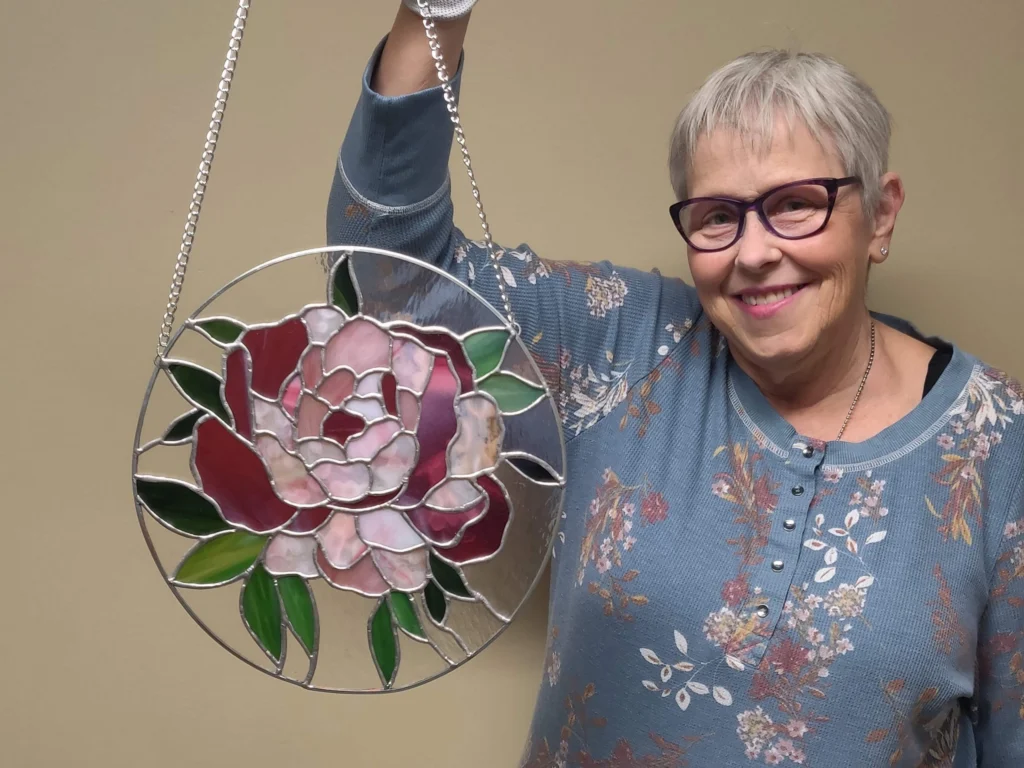 Woman holding a circular stained glass suncatcher featuring a pink and red peony with green leaves.