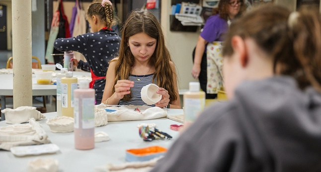 A young girl focuses on shaping a small white ceramic bowl during an arts and crafts class.