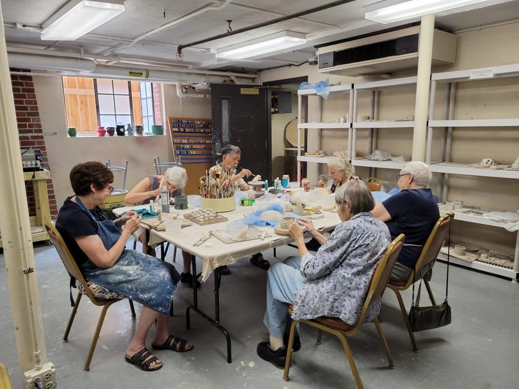 Seniors engaged in a pottery class, working with clay at a table in a well-lit studio.