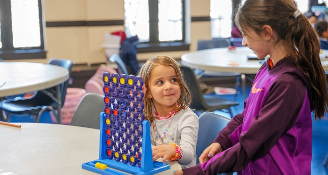 Two young girls playing Connect Four at a table in a room with round tables and chairs.