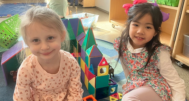 Two young girls smile while sitting next to a colorful magnetic block structure.