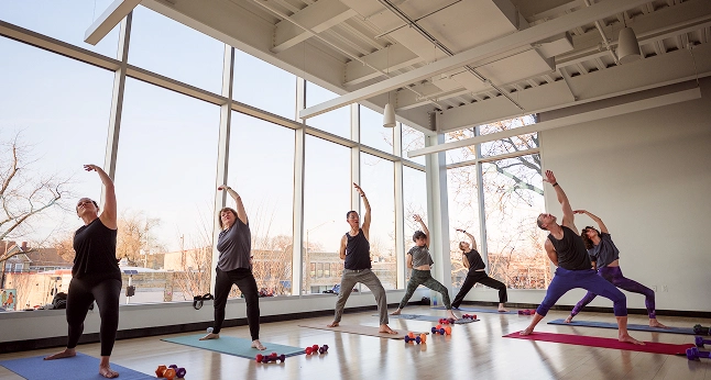 A diverse group of people practicing yoga in a bright studio with large windows. They are performing various stretching
