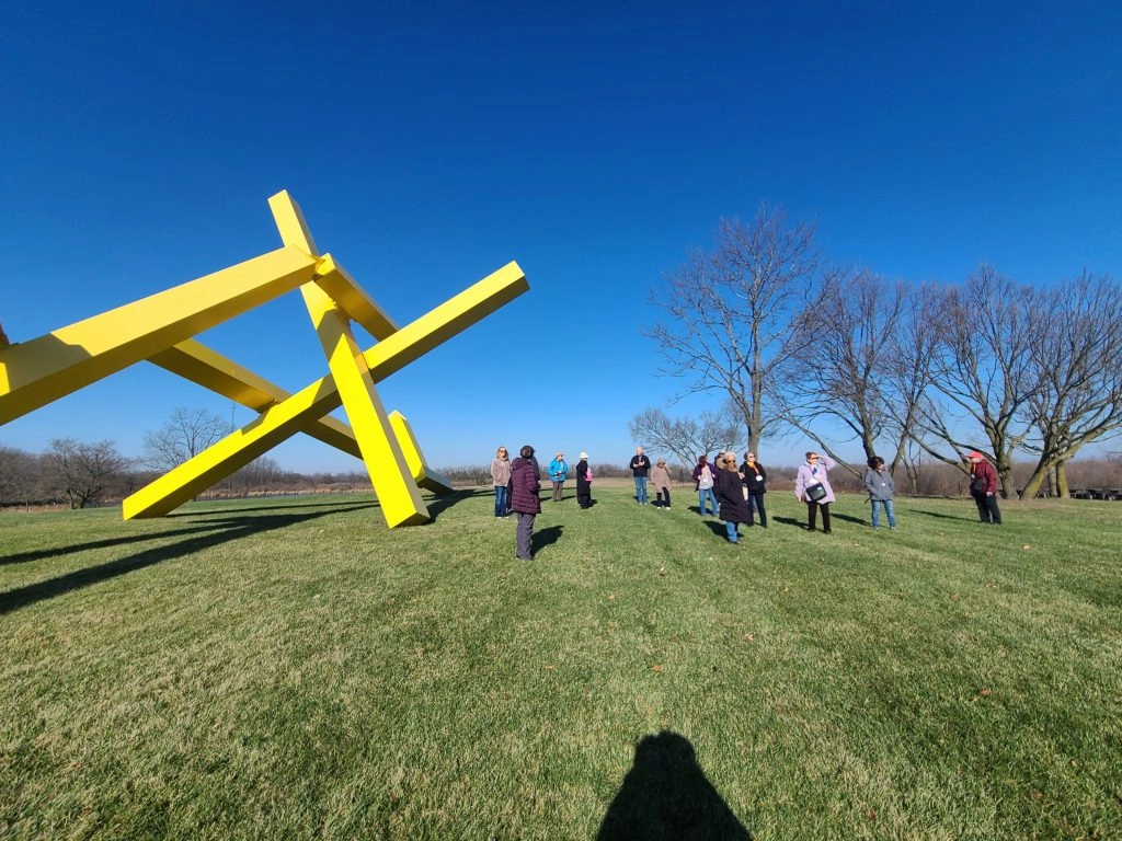 A group of people observe a large, bright yellow abstract sculpture on a grassy hill under a clear blue sky.