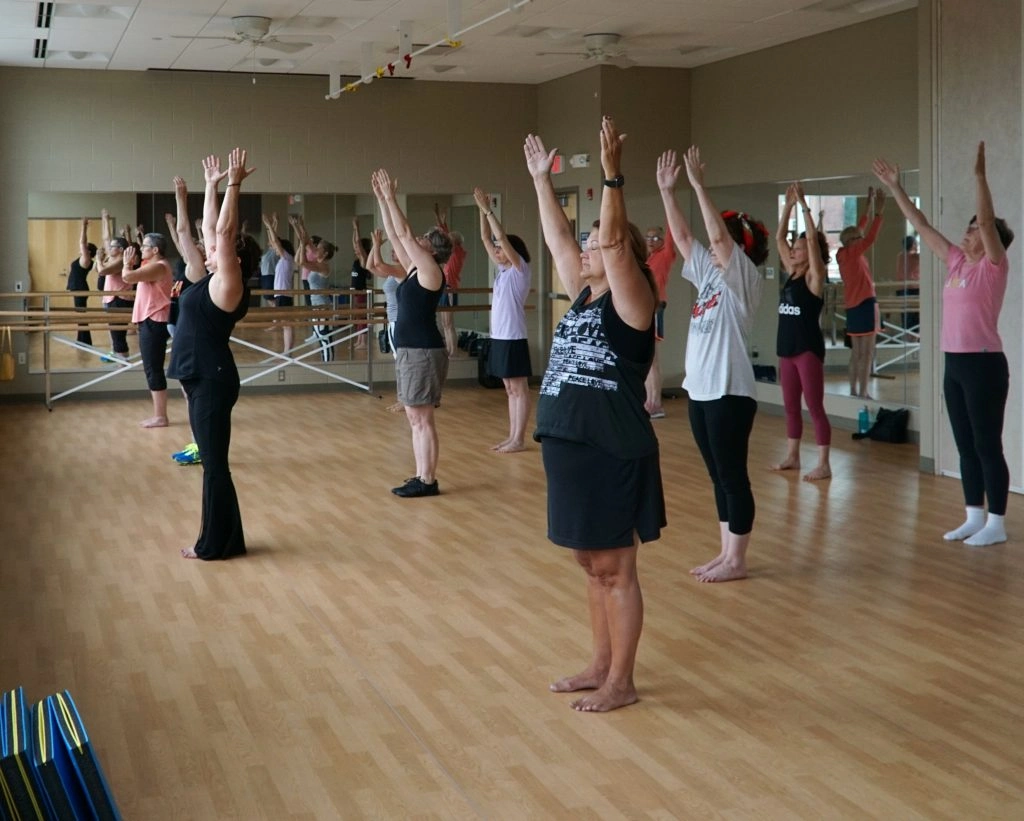 A diverse group of adults participate in a fitness class, with arms raised overhead in unison.