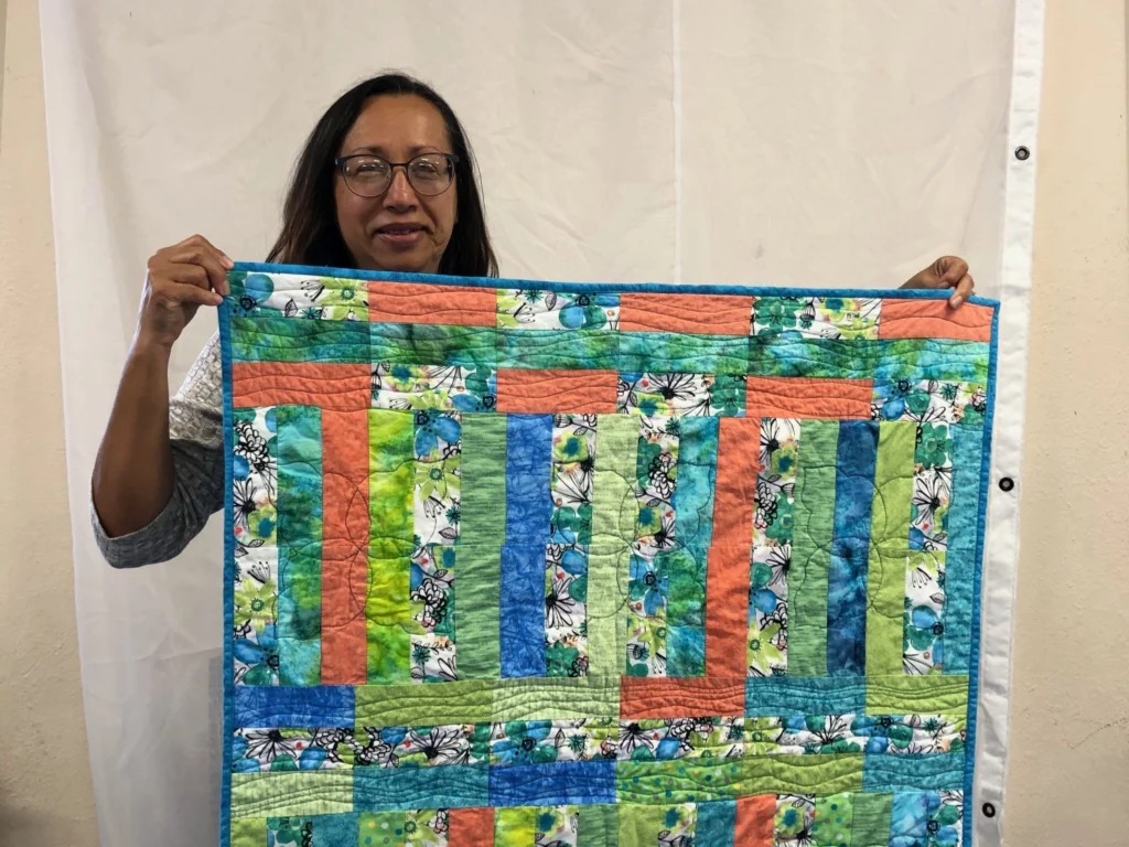 Woman holding a colorful quilt with green, blue, and coral fabric strips and floral patterns.