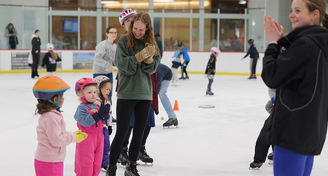 A group of children and adults on an ice rink, some wearing helmets and skates. A woman in a green sweatshirt claps her