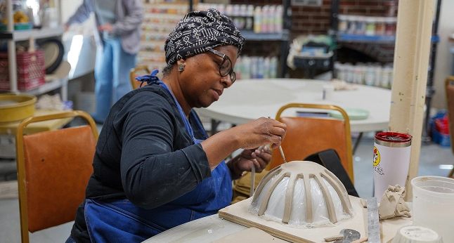 Woman wearing a headwrap and blue apron works on a clay bowl, using a tool to shape the rim.