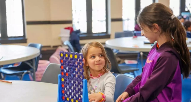 Two young girls play a Connect Four game at a table in a bright room.