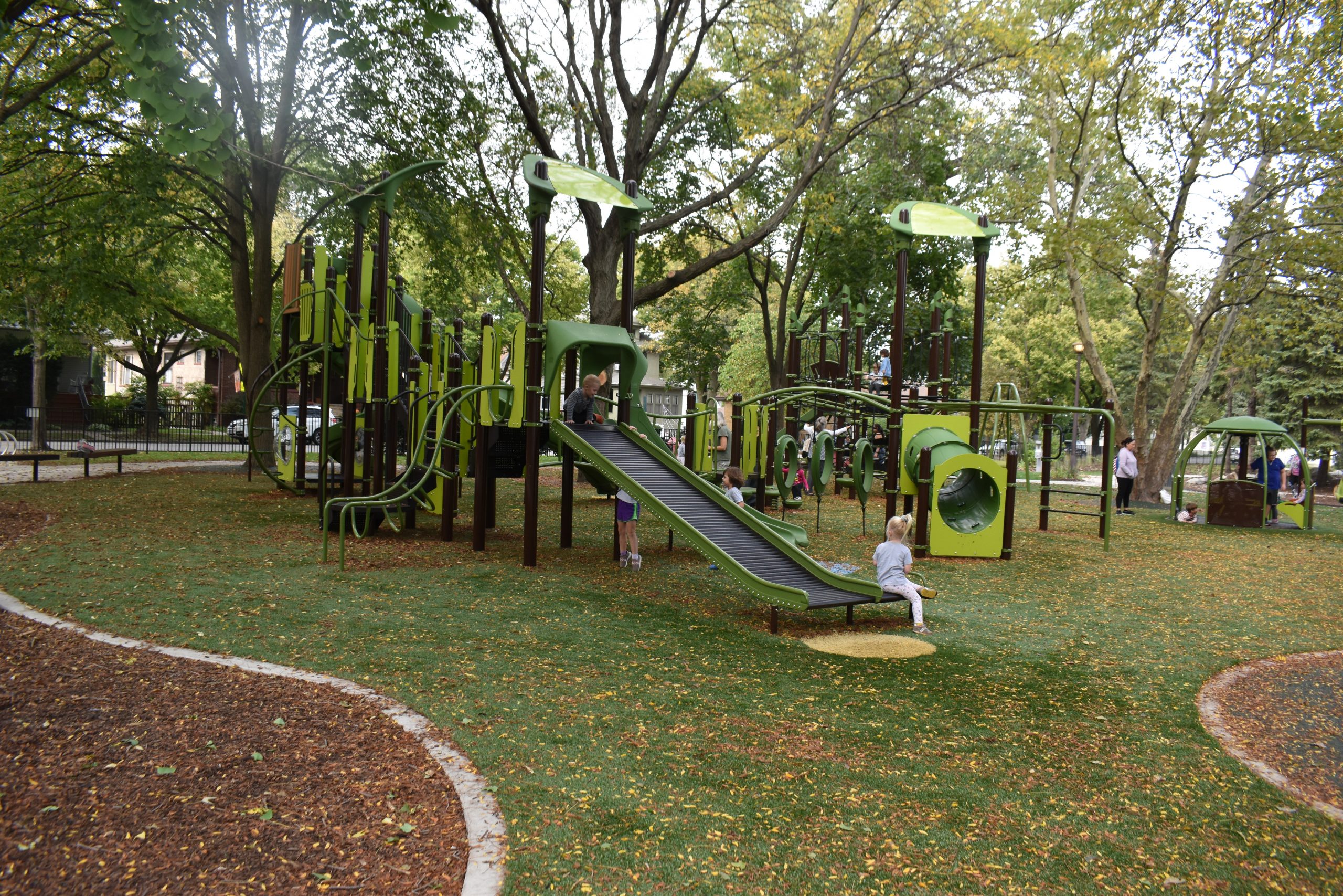 A modern green and brown playground with slides, tunnels, and climbing structures. Children are playing on the equipment.