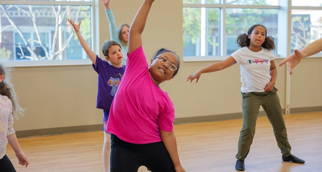 Children in a dance class practicing movements with their arms raised and bodies bent.