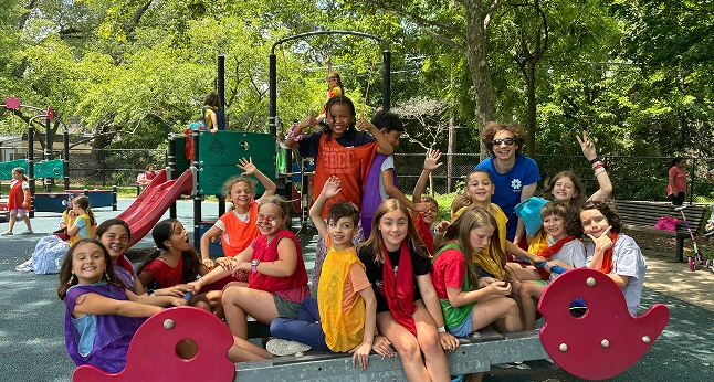 A diverse group of happy children and a counselor pose for a photo on playground equipment during a summer camp.