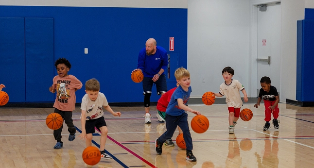 A coach instructs young children dribbling basketballs on a court. Several children are actively engaged in the drill.