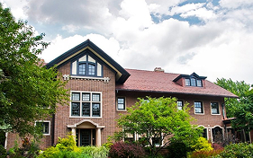 Cheney Mansion, a large brick building with a red tile roof, surrounded by lush greenery and trees under a cloudy sky.