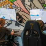 Two children play video games on curved monitors in an esports setup, with colorful keyboard-themed murals on the wall.