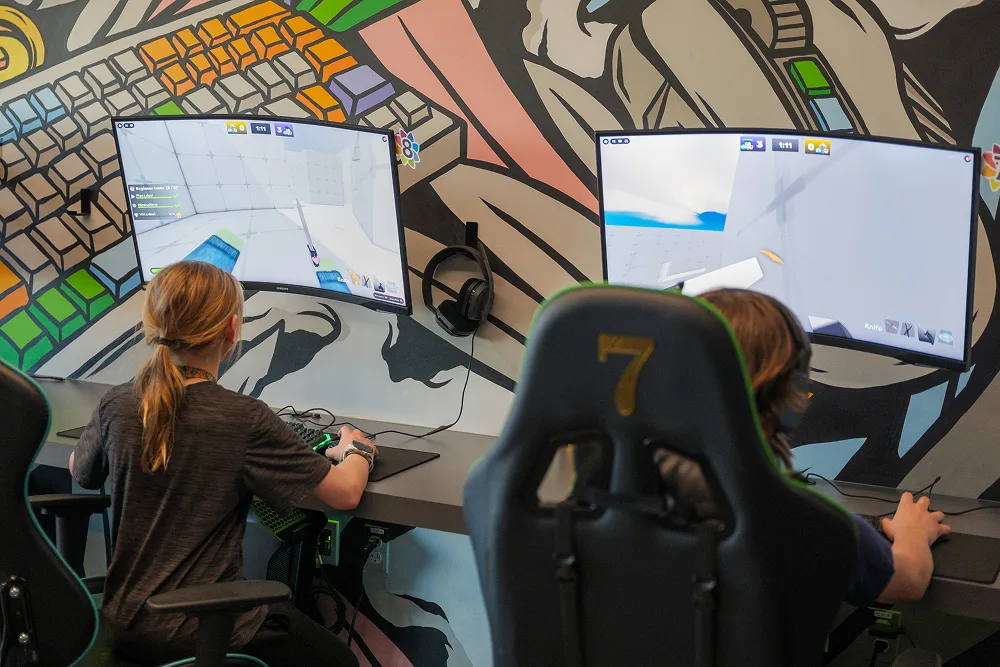 Two children play video games on curved monitors in an esports setup, with colorful keyboard-themed murals on the wall.