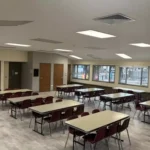 Rectangular tables with burgundy chairs around them set up in a spacious room with wood doors and large windows.