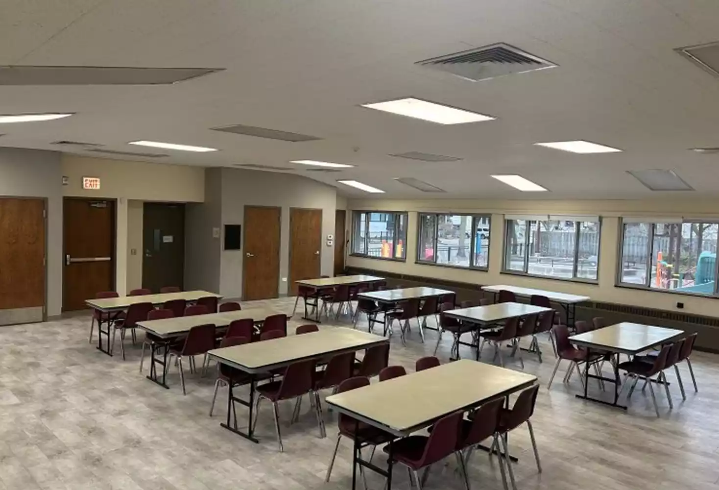 Rectangular tables with burgundy chairs around them set up in a spacious room with wood doors and large windows.