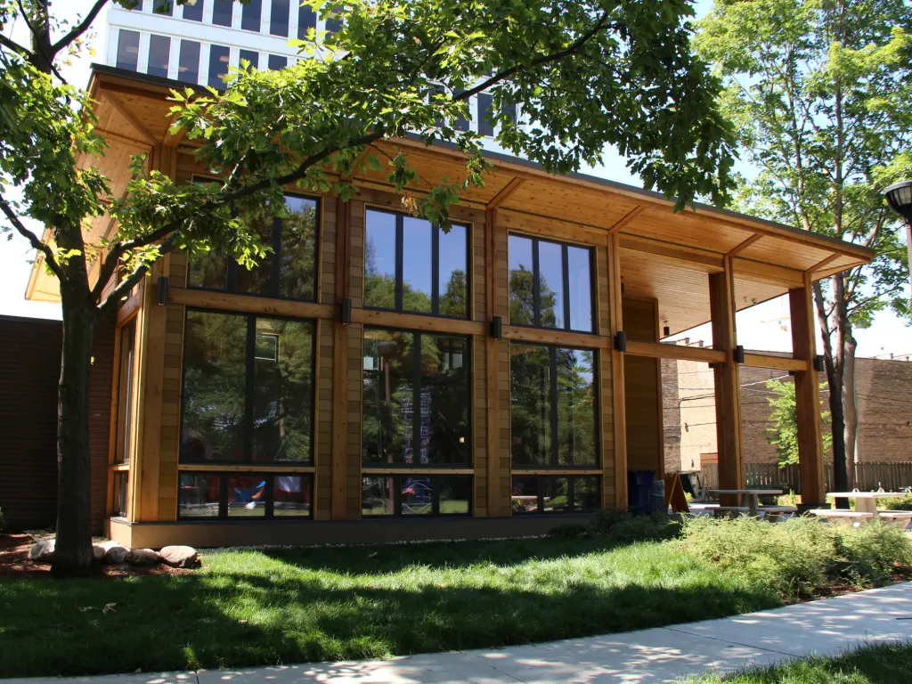 Modern wooden building with large windows and an overhang, surrounded by trees and green lawn.