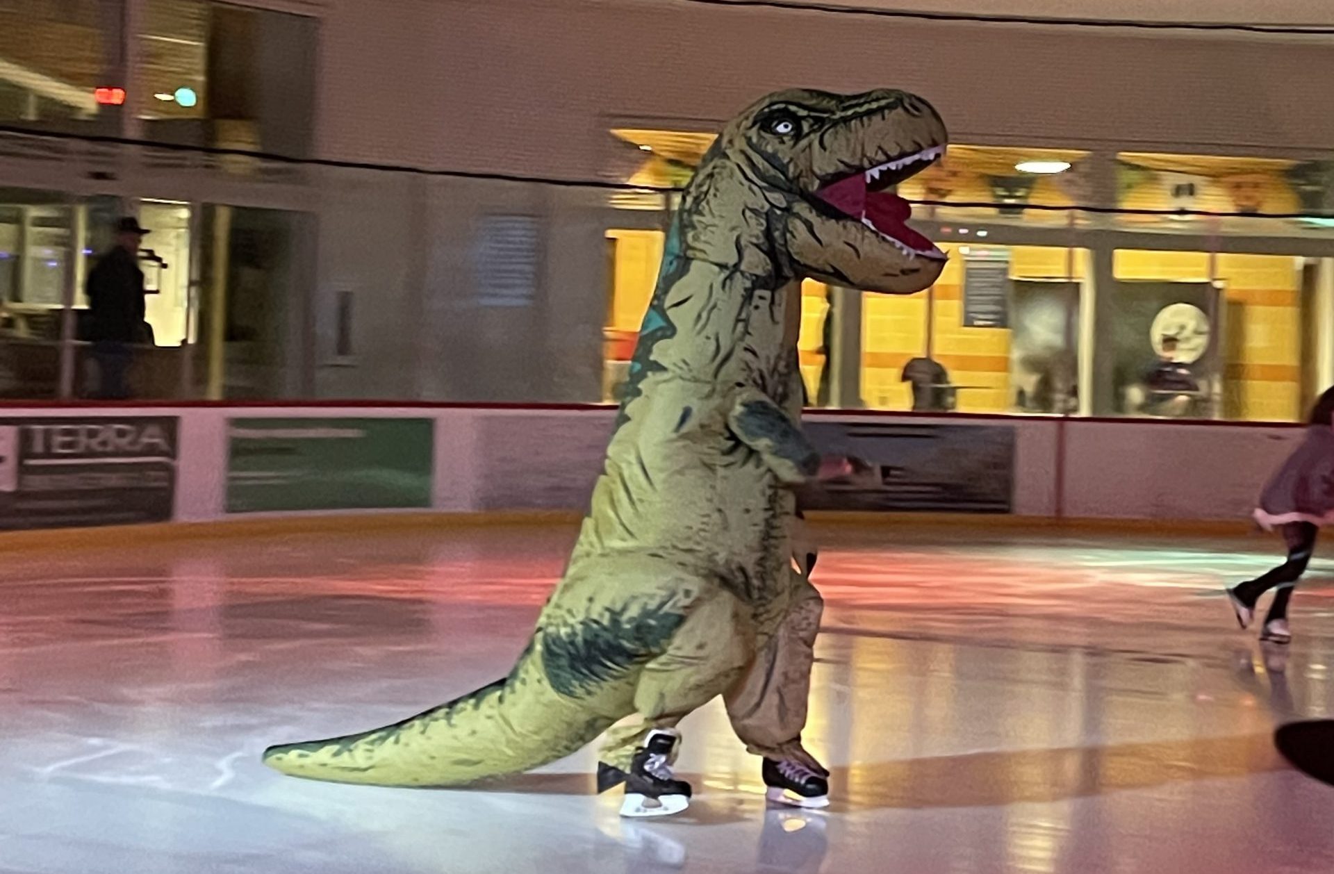 A person in a T-Rex costume skates on an ice rink during an ice skating party.