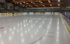 An empty indoor ice rink with spectator seating visible in the background. The ice surface is smooth and reflects the