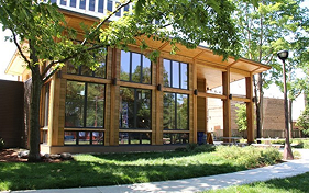 Modern wooden building with large windows, surrounded by green grass and trees, suggesting a sustainable garden space.