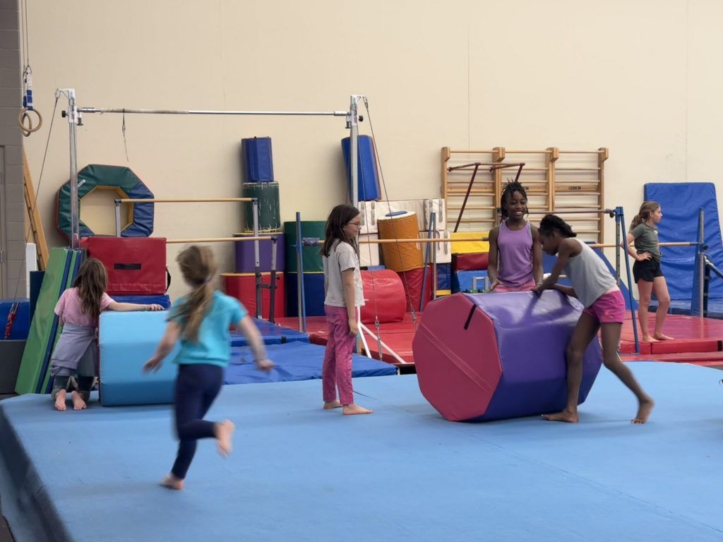 Girls practice gymnastics, with some using a purple barrel and others near bars and mats.