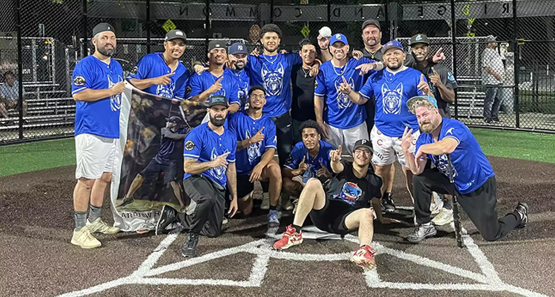 A baseball team in blue jerseys with a wolf logo poses for a photo on a baseball field, holding a banner.