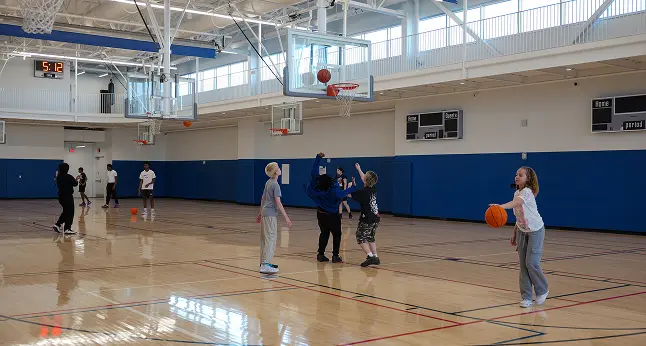 Teens play basketball in an indoor gymnasium. A girl dribbles a ball, while others practice shooting and passing.