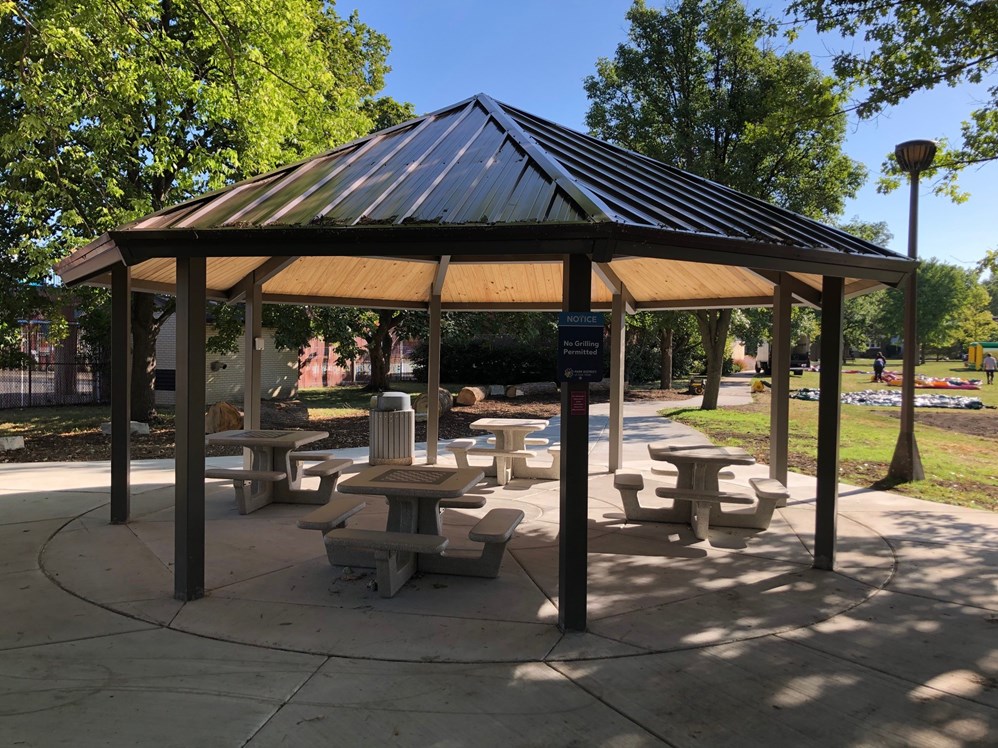Octagonal gazebo with a metal roof, wooden interior ceiling, and concrete picnic tables. A 'No Grilling Permitted' sign is