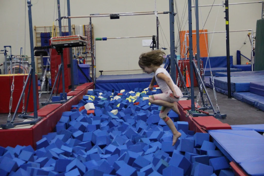 A young girl in athletic wear jumps into a foam pit filled with blue, yellow, and red foam cubes at a gymnastics center.