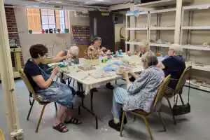 Several adults sit around a table, engaged in a craft activity. They appear to be working on a project with yarn or thread.
